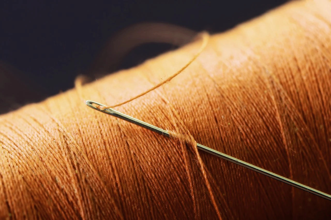 Close-up of a silver needle threading through copper-colored thread on a spool. The focus highlights the intricate fibers, conveying precision and craftsmanship.