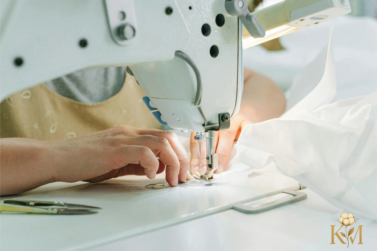 A person sewing fabric on a machine, focused on precise stitching, with scissors nearby and soft fabric draped around.