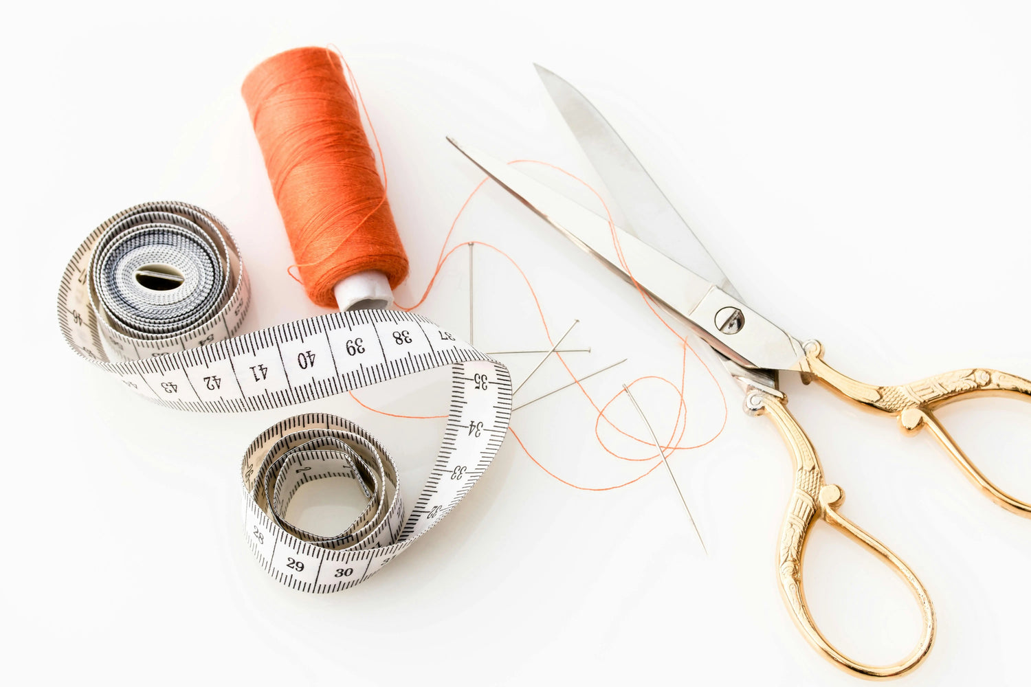 A spool of orange thread, measuring tape, scissors, and sewing needles on a white background, arranged for sewing projects.
