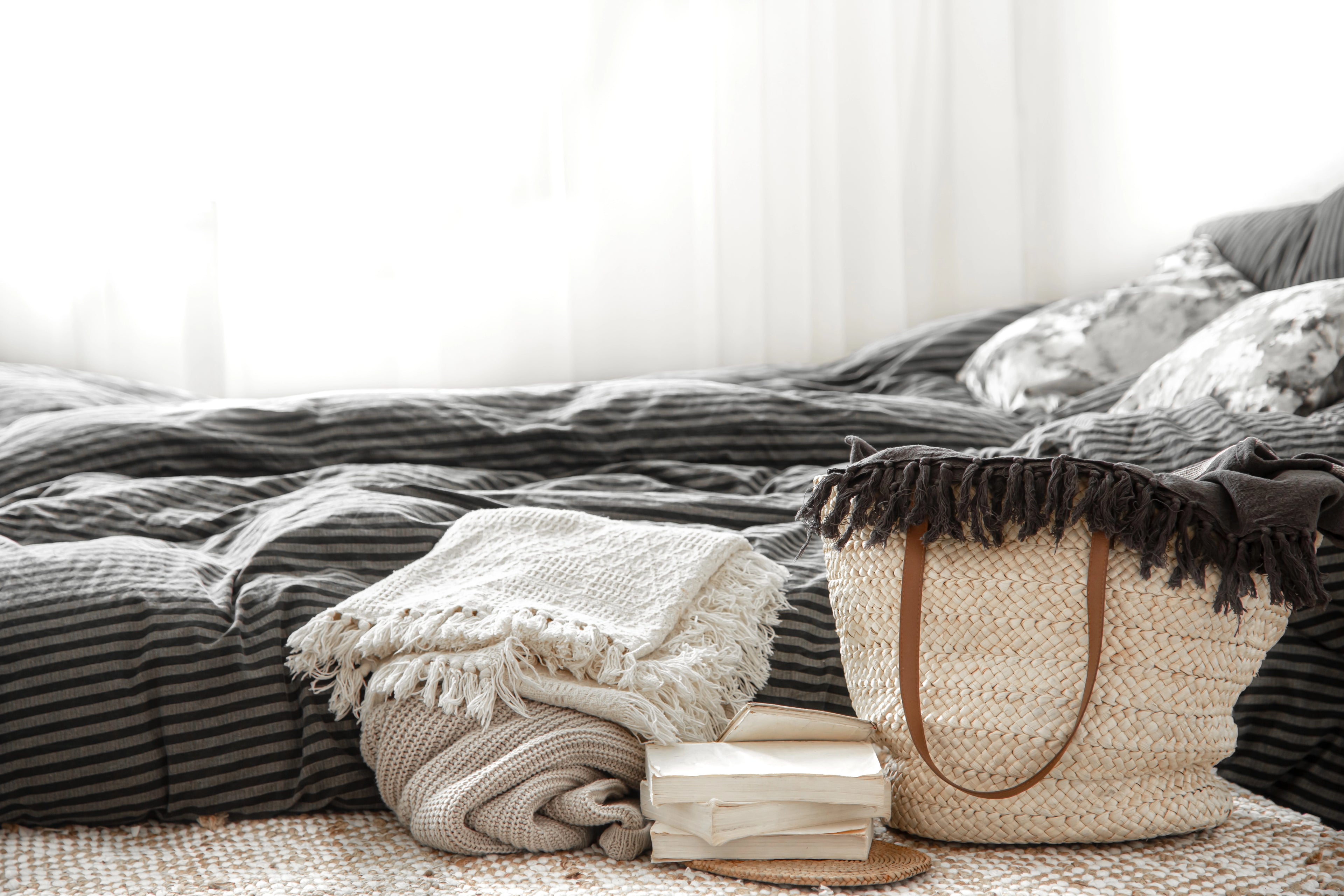 A neatly made bed with dark striped bedding is in the background. In the foreground are folded blankets, two books, and a woven straw bag with leather handles.